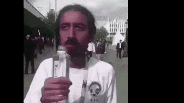 a man with a beard is drinking water from a bottle while wearing a ksa shirt .