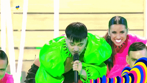 a man in a neon green jacket is singing into a microphone while a group of dancers are behind him .