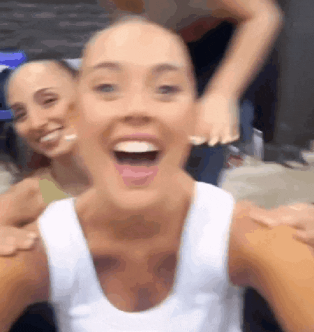 a woman in a white tank top is laughing with her mouth open while another woman looks on .