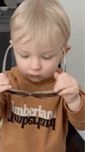 a young boy wearing a timberland shirt is holding a pair of sunglasses in his hands .
