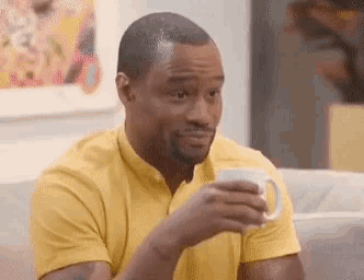 a man in a yellow shirt is sitting on a couch drinking from a cup .