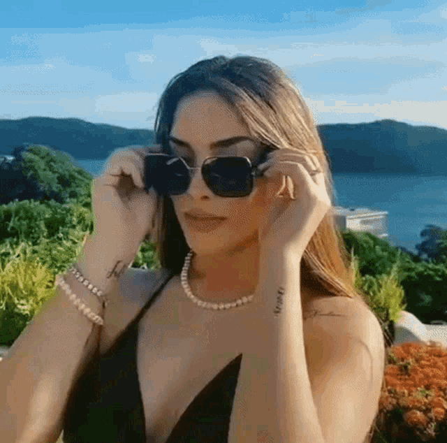 a woman wearing sunglasses and a pearl necklace is standing in front of a lake .