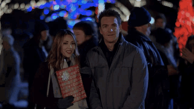 a man and a woman standing next to each other holding a red gift bag that says merry christmas