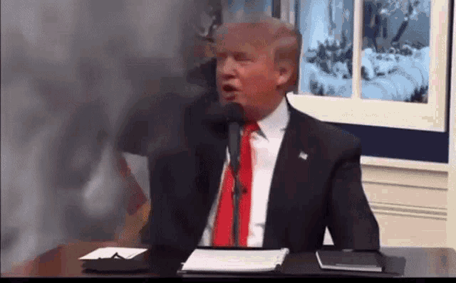 donald trump is sitting at a desk talking into a microphone while wearing a suit and tie .