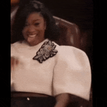 a woman is sitting in a chair and smiling . she is wearing a white top with puffed sleeves .