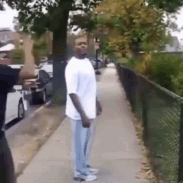 a man in a white shirt and blue pants is standing on a sidewalk next to a fence .