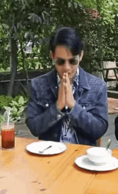 a man in a denim jacket sits at a table with his hands folded