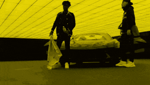 two men standing in front of a car with one wearing a black jacket with the letter m on the front
