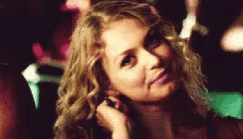a woman with blonde curly hair is sitting in a dark room and looking at the camera .