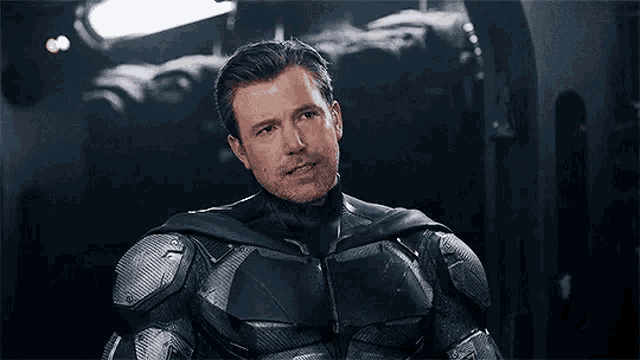 a man in a batman suit is sitting in a dark room and pointing at the camera .