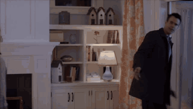 a man in a black coat stands in a living room with a lamp