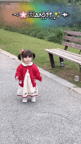 a little girl in a red cardigan and white dress stands in front of a wooden bench