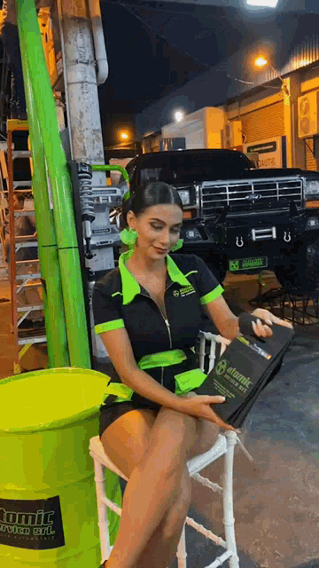 a woman sits on a chair next to a green barrel that says atomic