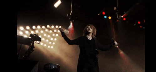 a woman in a black coat stands on a stage holding a bottle of water