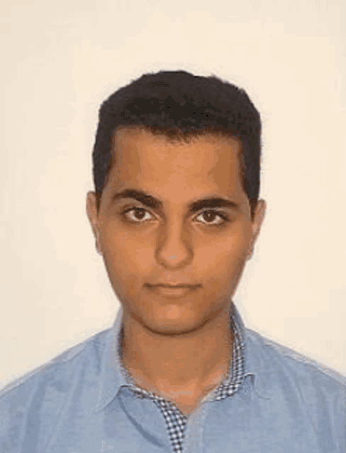 a young man in a blue shirt is standing in front of a white wall and looking at the camera .