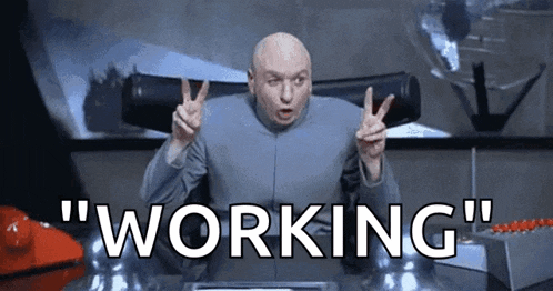 dr. evil is giving a peace sign while sitting at a desk and the words `` working '' are behind him .