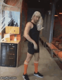 a man wearing a wig and orange socks is dancing in a room with a fridge in the background
