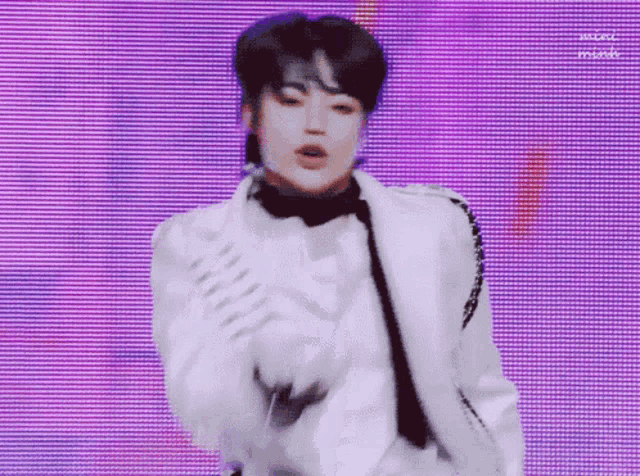 a young man in a white jacket and tie is dancing on a stage in front of a purple background .