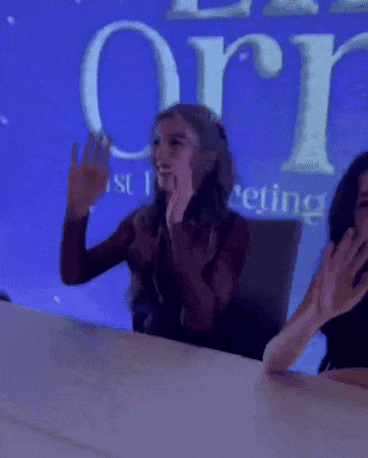 two women are sitting at a table with their hands in the air in front of a sign that says orion .