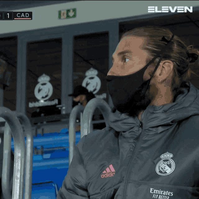 a man wearing a mask and an adidas jacket sits in a stadium