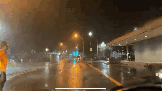 a man in an orange jacket stands on the side of a wet street at night