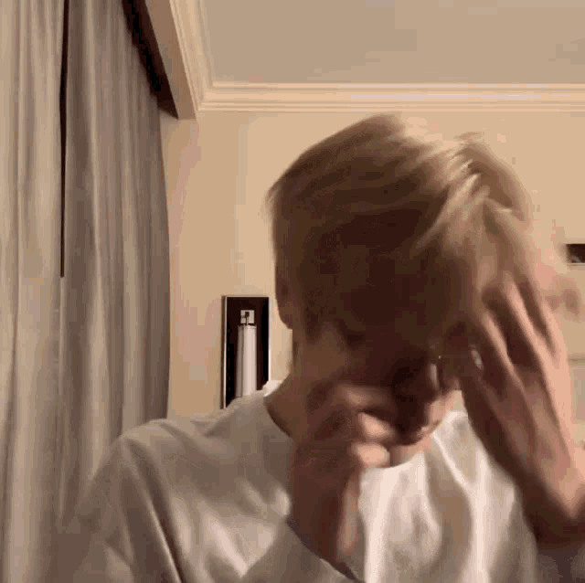 a man with blonde hair is covering his face with his hands in a room .