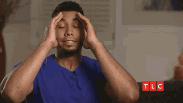 a man in a blue shirt is sitting on a couch holding his head .