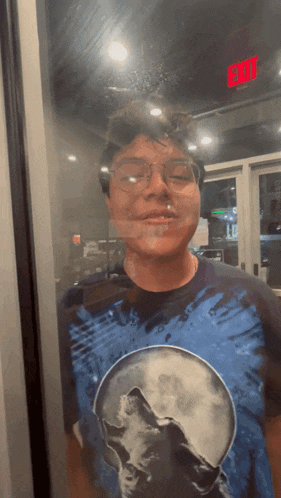a man wearing glasses and a blue shirt with a wolf on it stands in front of an exit sign