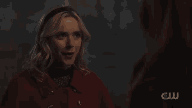 a woman in a red coat stands next to another woman with a cw logo on the bottom right