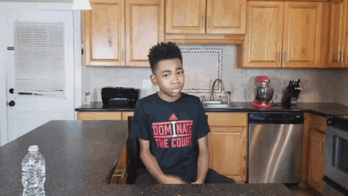 a boy wearing a black shirt that says dominate the court sits in a kitchen