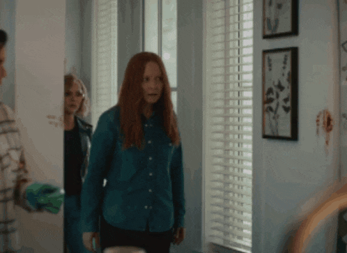 a woman with red hair stands in front of a window with blinds