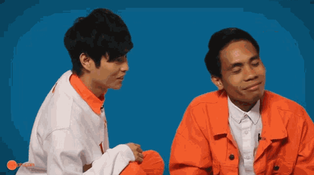 two men in orange jackets are sitting next to each other on a blue background with a smiley face on the bottom right corner