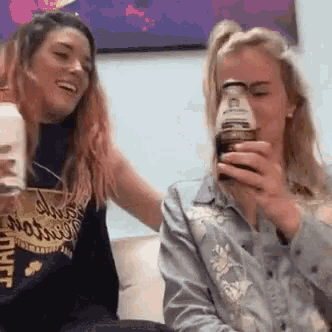 two women are sitting on a couch laughing and drinking from a bottle .