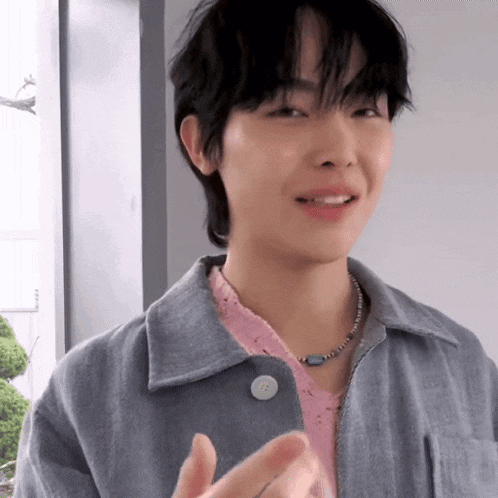 a young man wearing a grey jacket and a pink shirt is making a heart with his hands .