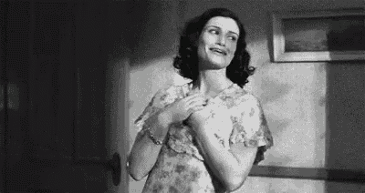a black and white photo of a woman in a dress standing in a room with her hands on her chest .