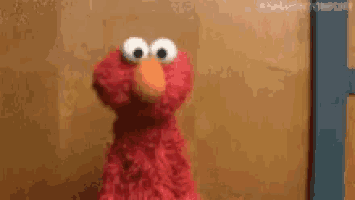 elmo from sesame street is standing in front of a brown wall and looking at the camera .