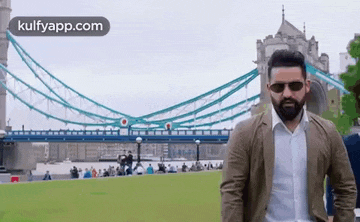 a man wearing sunglasses and a suit is standing in front of a bridge .