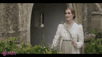 a woman in a white coat is holding a flower in front of a stone wall