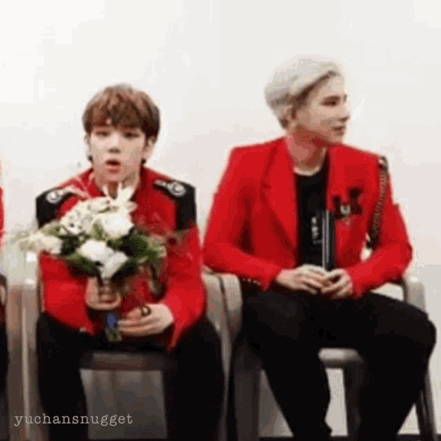 a man in a red jacket is holding a bouquet of flowers while sitting next to another man in a chair .