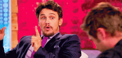 a man in a suit and tie is pointing at another man while sitting in front of a pink wall .