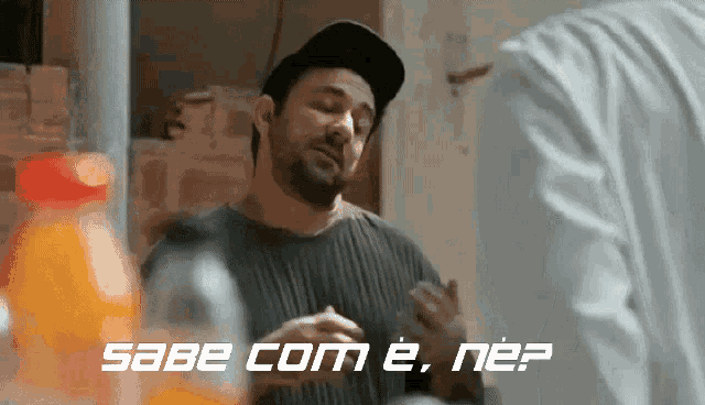 a man wearing a hat talks to another man with the words sabe com e. nee written in white