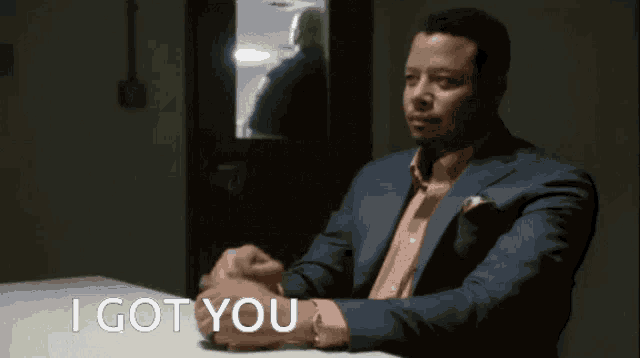 a man in a suit sits at a table with the words " i got you " behind him