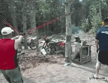 two men are playing a game of slingshot in the woods with motorcycles in the background .