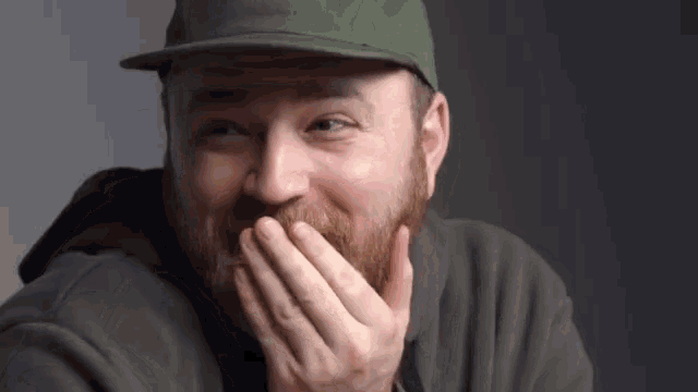 a man with a beard wearing a hat is covering his mouth with his hands .