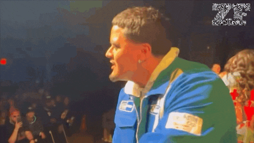 a man in a blue and green jacket stands in front of a crowd with the number 20 on the bottom