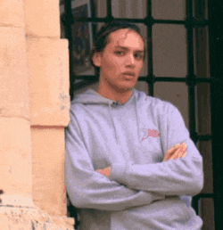 a man leaning against a wall with his arms crossed wearing a gray sweatshirt