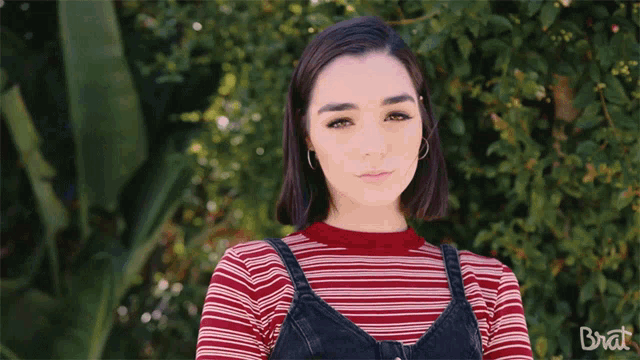 a woman wearing a red and white striped shirt and overalls stands in front of a tree with the word brat on the bottom