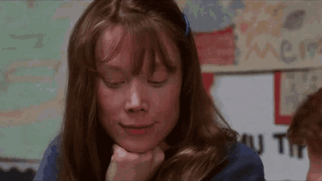 a girl with long brown hair is sitting in a classroom with her eyes closed and her hand on her chin .