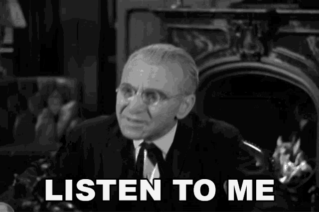 a man in a suit and tie is sitting in front of a fireplace and a sign that says listen to me