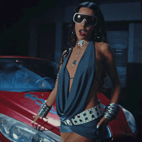 a woman wearing sunglasses and a blue dress is standing next to a red car